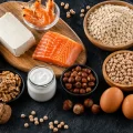 Overhead view of high-protein foods including salmon, chicken breast, eggs, tofu, shrimp, yogurt, oats, chickpeas, lentils, beans, walnuts, and hazelnuts on a dark background.