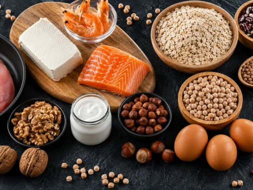 Overhead view of high-protein foods including salmon, chicken breast, eggs, tofu, shrimp, yogurt, oats, chickpeas, lentils, beans, walnuts, and hazelnuts on a dark background.